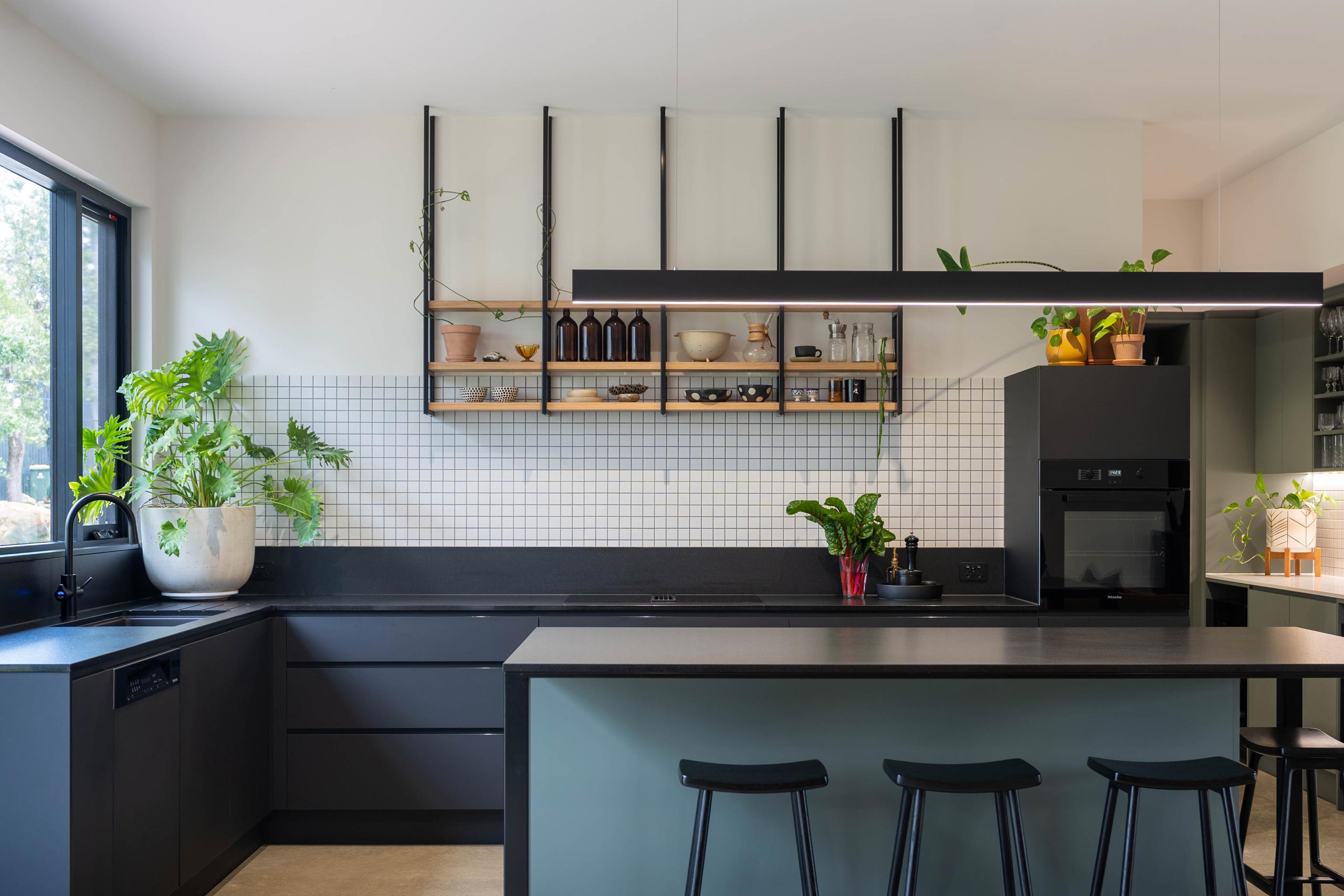 A Natural, industrial beauty within a kitchen. Warm tones with teal and black touch cabinetry, a white tiled square splashback, black slim benchtop and open shelving. above.