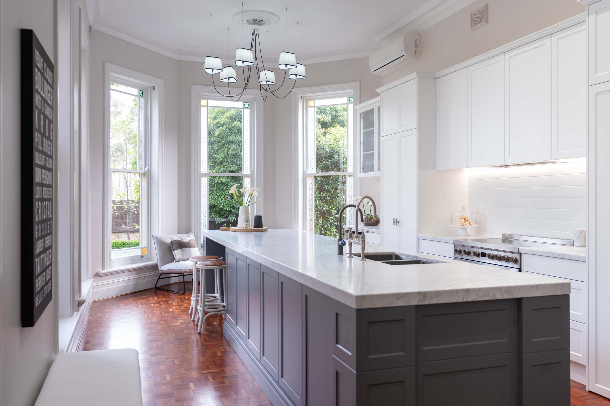 In a setting of grandeur, the Hamptons style kitchen is perfectly harmonious amongst traditional elements of parquetry floors, high ceilings with ornate cornice, and adorned windows opening out onto the lush green garden beyond.