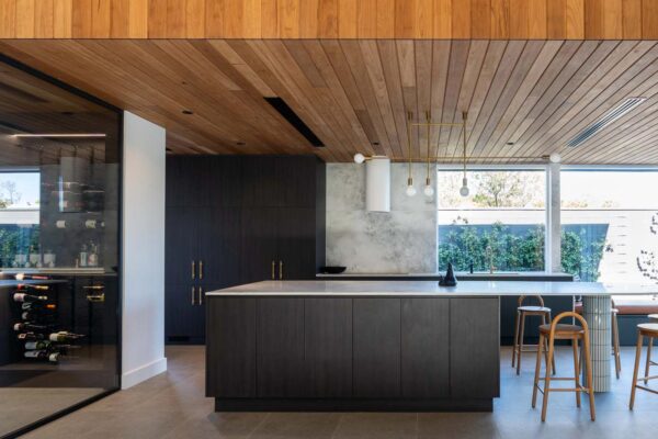 An open plan kitchen with adjacent glass wine room with a curved Kitchen Island in Laminex burnished wood, a deep warm brown, complimenting an elegant marble benchtop and kit-kat tile column support with a wood panel ceiling bulk head.