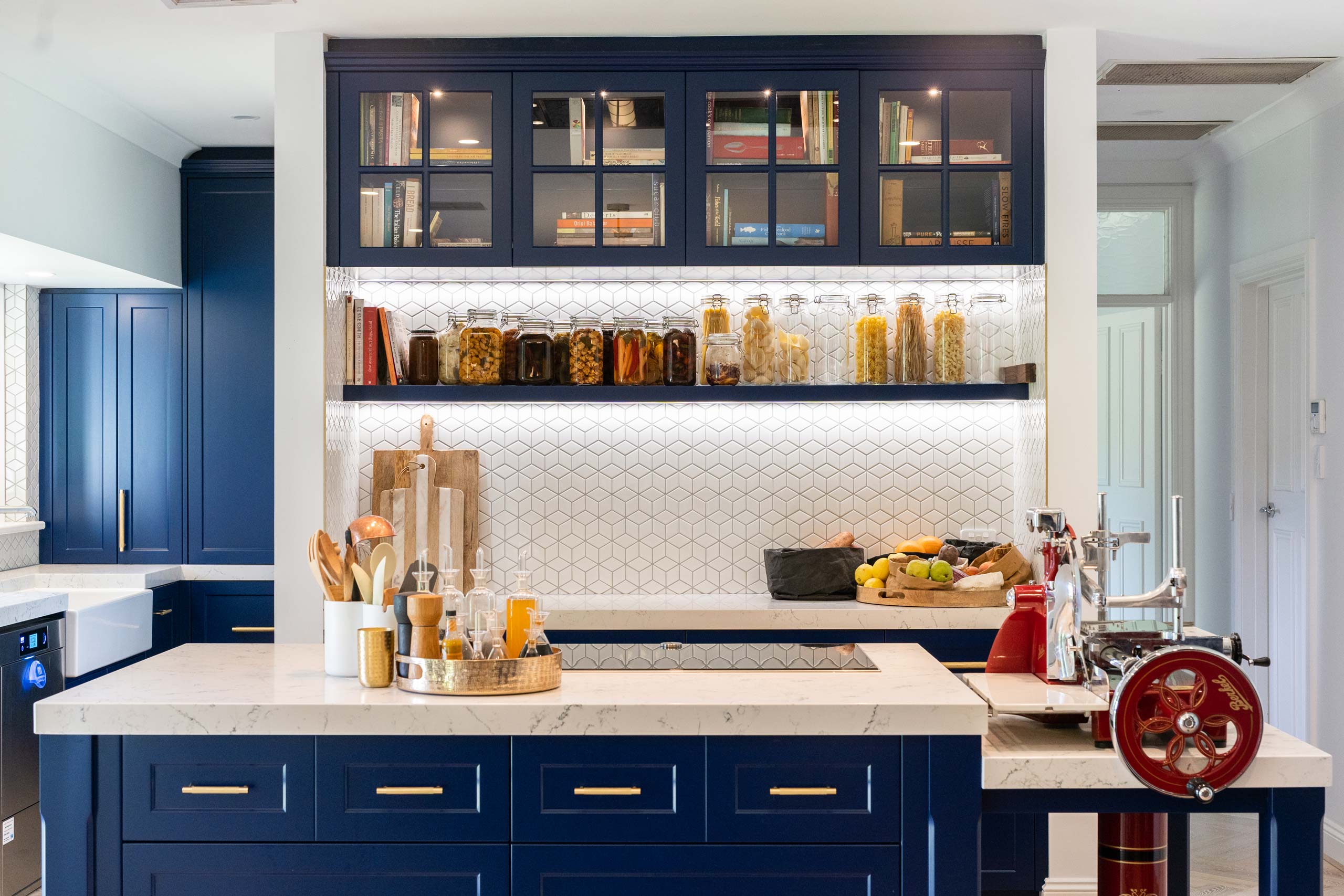 European, bold Navy kitchen with glass storage and shelving spaces. A white diamond tiled splashback and hidden pantry space, creates a versatile kitchen space for cooking.