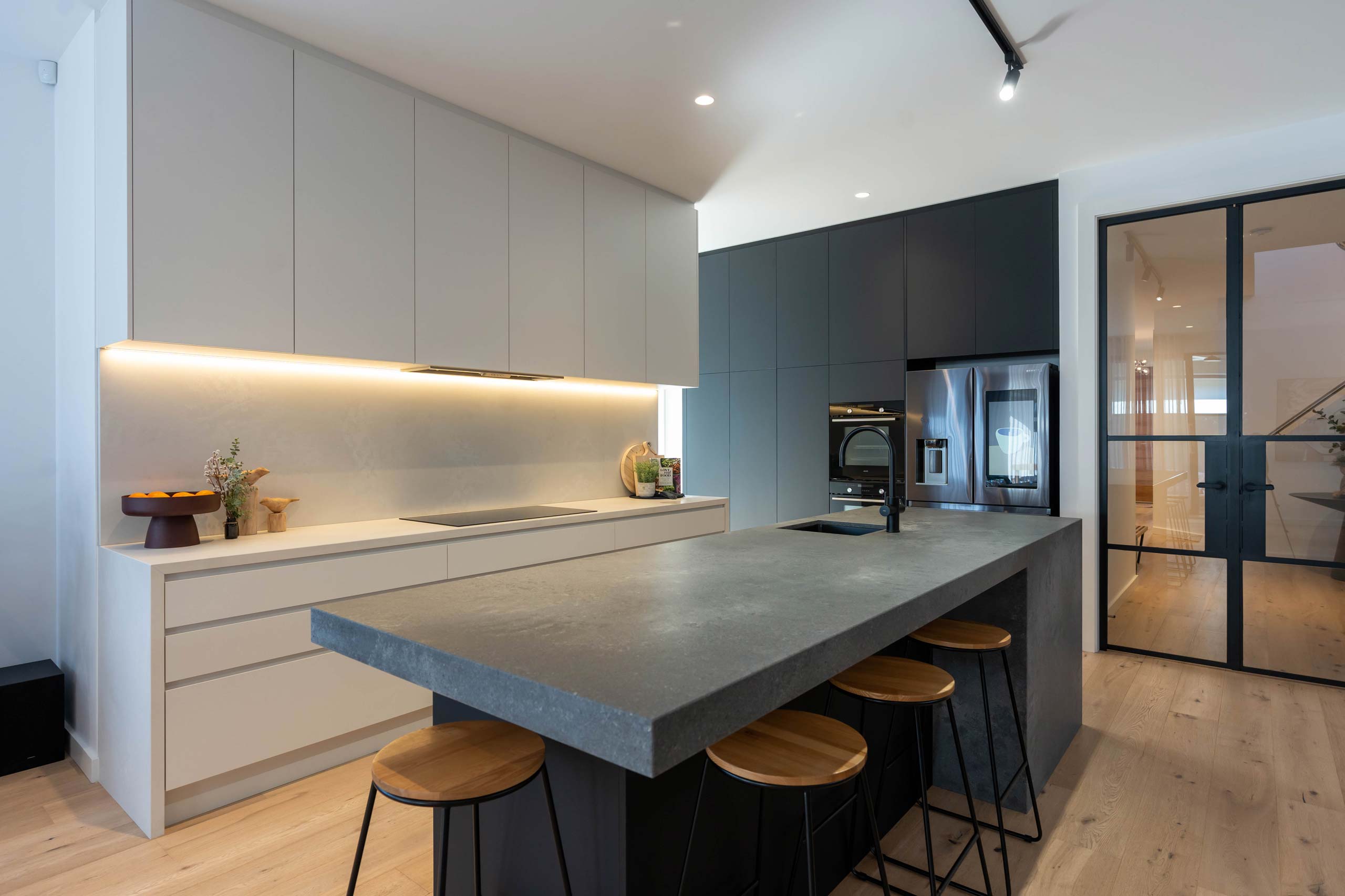 Black and white Kitchen. Island bench concrete look stone, wooden stools and black tapware. Contrasting white galley with ceiling height overheads.