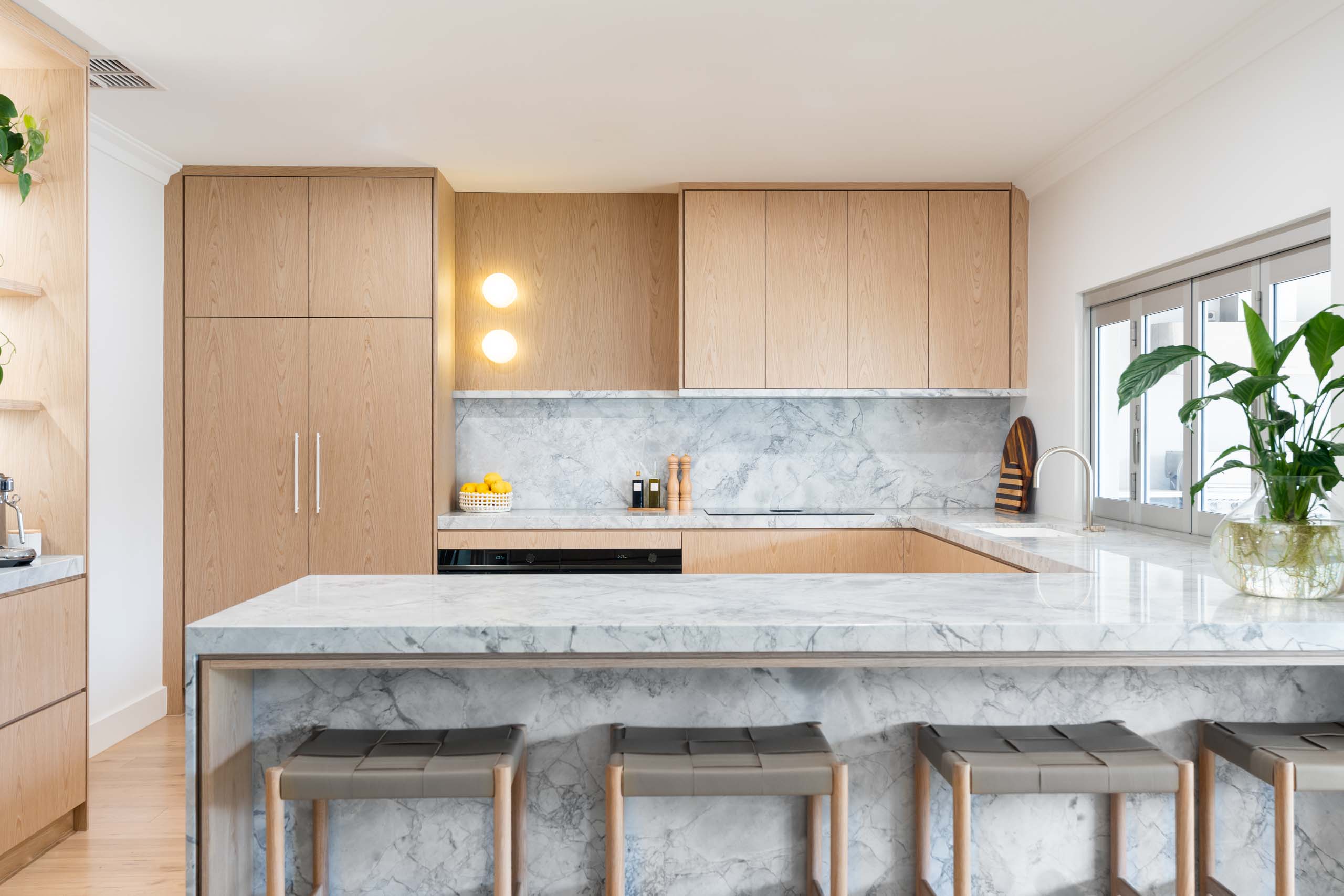 A luxurious blond timber veneer kitchen with a delicate marble. This kitchen is highlighted by the marble waterfall benchtop and open shelving with a stone base.