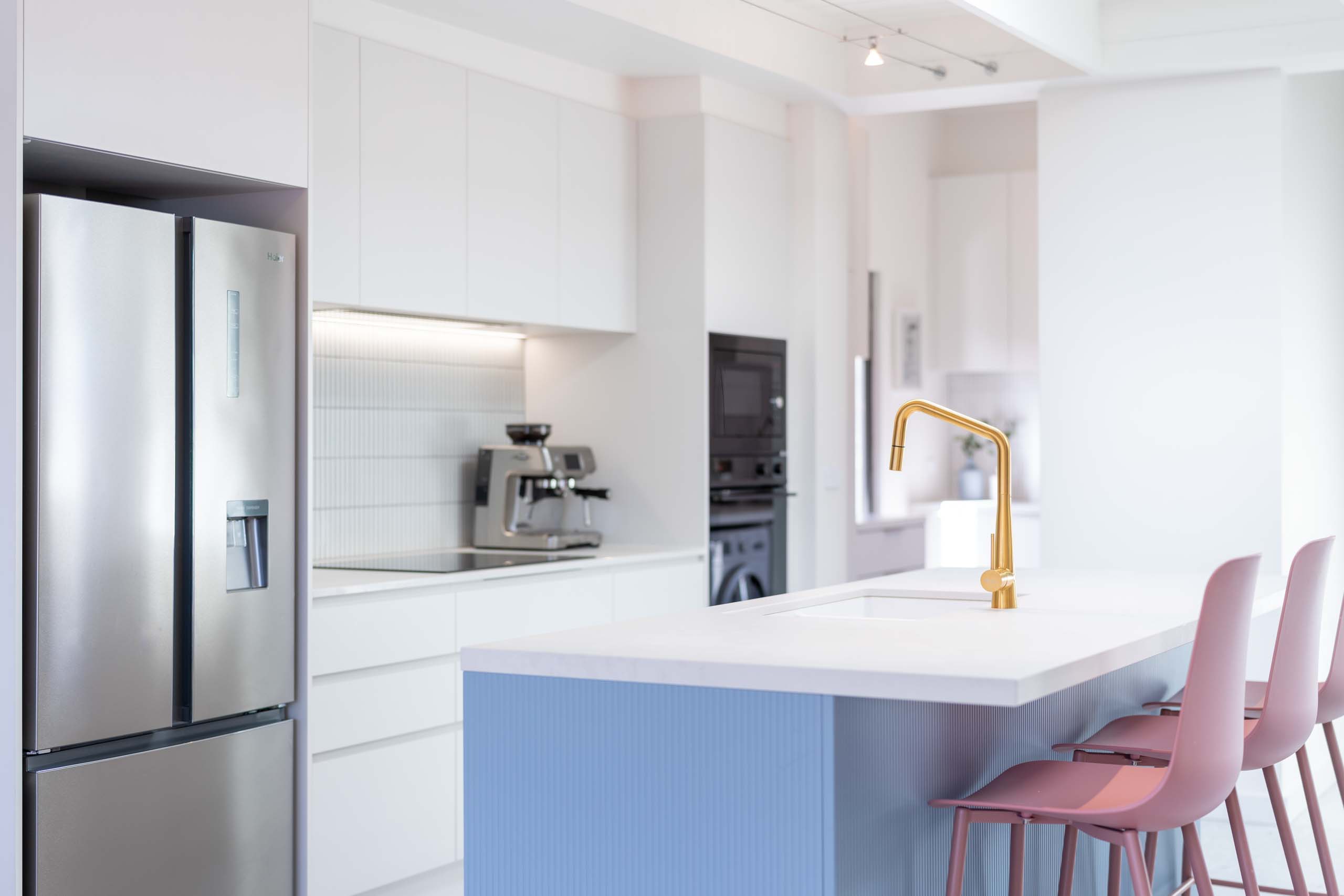 White kitchen with blue denim island bar and pink stools. Gold Tap and wall oven.