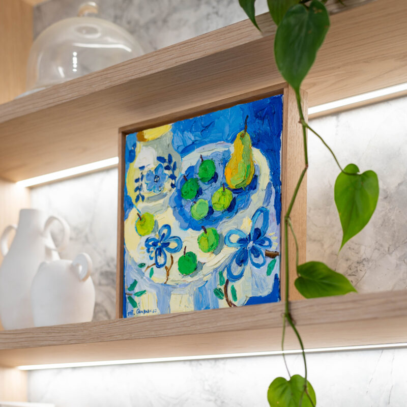 Kitchen delicately organic. Blond timber veneer, natural marble, shelving with bright blue art work.