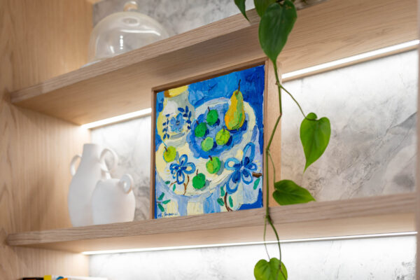 Kitchen delicately organic. Blond timber veneer, natural marble, shelving with bright blue art work.