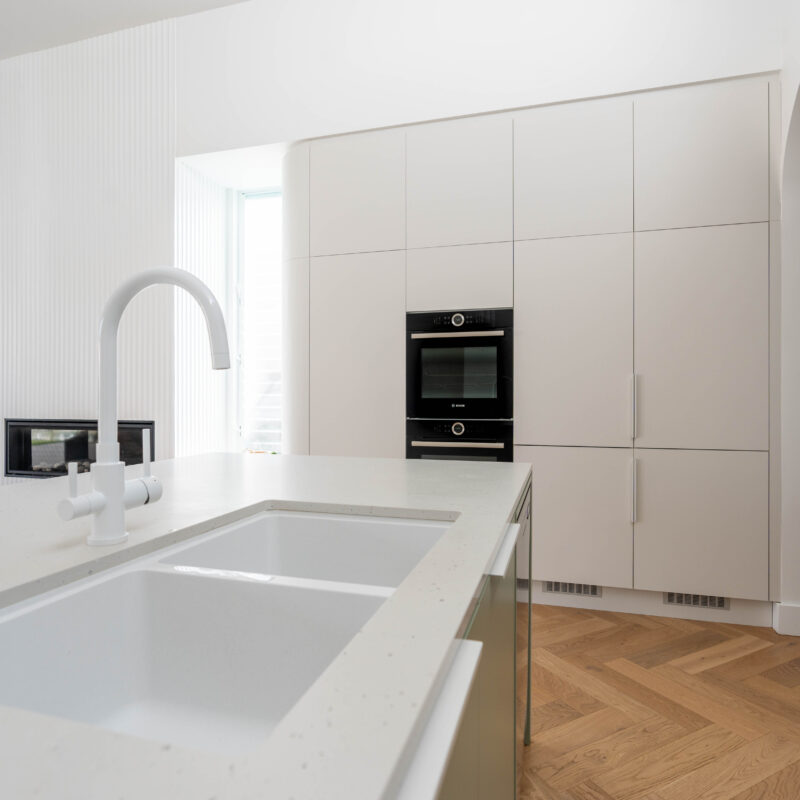 Kitchen. Calming curves. White grey curved cabinetry with oven wall and storage on a timber floor and white sink and tap