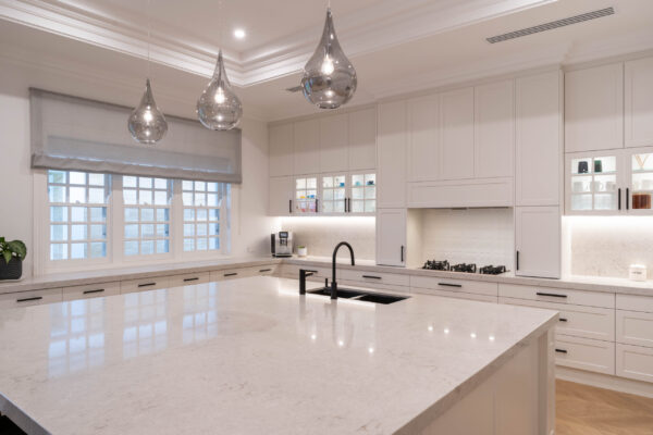 Hamptons Glamour. Classic shaker-style cabinetry and touches of modern glamour. The pearlescent finish of the cabinets in ghost gum satin swathes the space with the feel of luxury. The bold, black metal accents add a modern edge to the design with huge stone entertaining island bar.