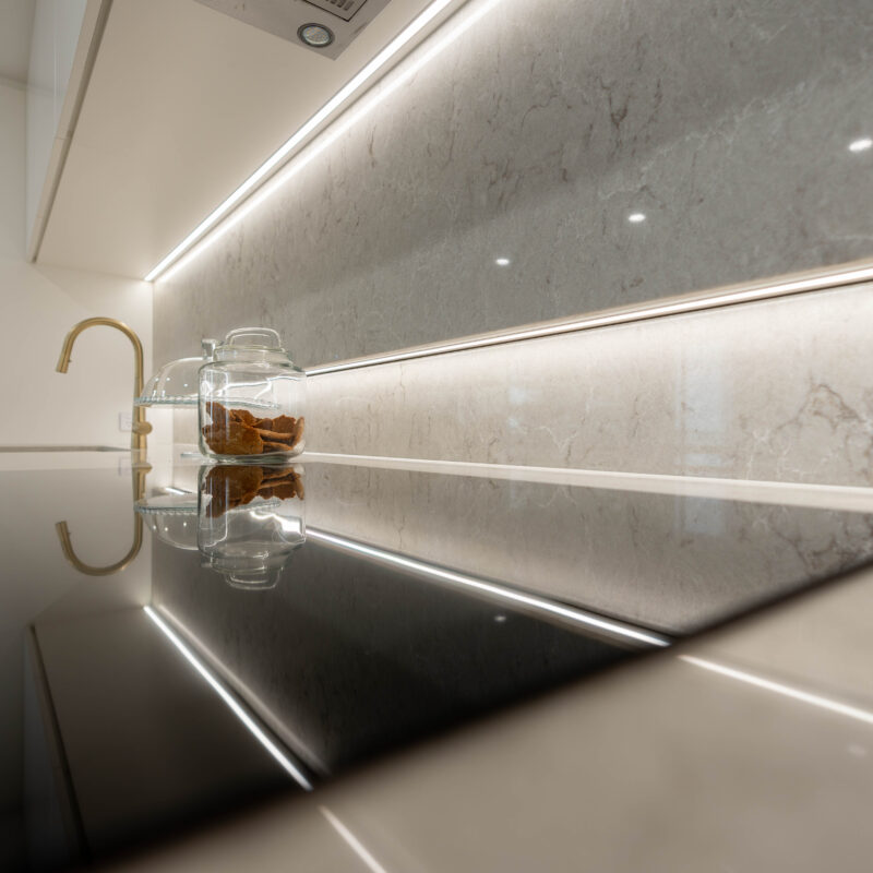 Illuminated Cooking space with electric black cooktop and soft stone beige splashback and gold tap.