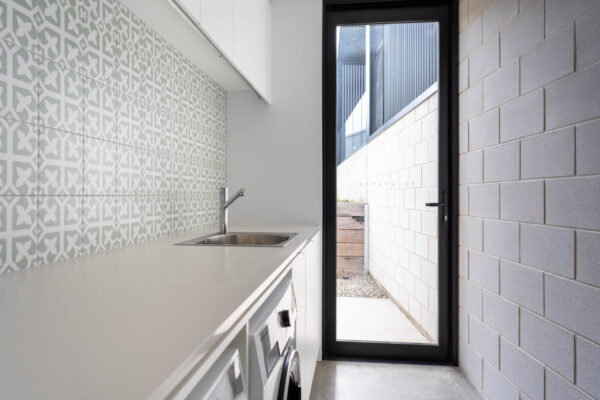 Long Laundry space with tub, chrome tap and green pattern splashback and black back door to the outside.