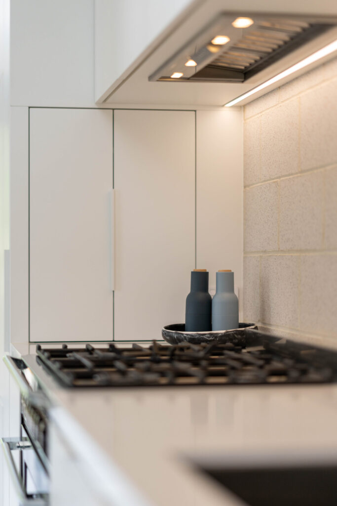 White cabinetry with natural breeze block splashback, cooktop and integrated rangehood and appliance nook.