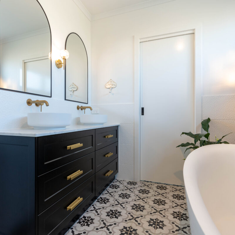 Bathroom Vanity- Double sink dark cabinetry with gold handles. Double mirrors and embossed white tiles splashback and patterned black, white and grey floor tiles with freestanding bath