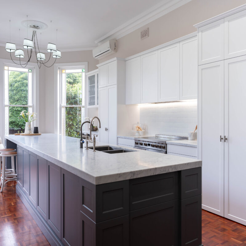 The mid tone neutral grey of the island provides a depth of colour between the light and bright white cabinetry, and traditional warm toned parquetry. Entertainer's Delight The core kitchen space is balanced and mirror image. Extending down towards the windows, the kitchen transforms into an entertaining space; with a glass door cabinet, wine fridge, and space for four stools, mimicking a traditional table setting, with two either side.