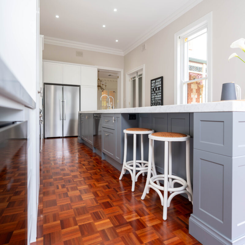 The mid tone neutral grey of the island provides a depth of colour between the light and bright white cabinetry, and traditional warm toned parquetry. Entertainer's Delight The core kitchen space is balanced and mirror image. Extending down towards the windows, the kitchen transforms into an entertaining space; with a glass door cabinet, wine fridge, and space for four stools, mimicking a traditional table setting, with two either side.