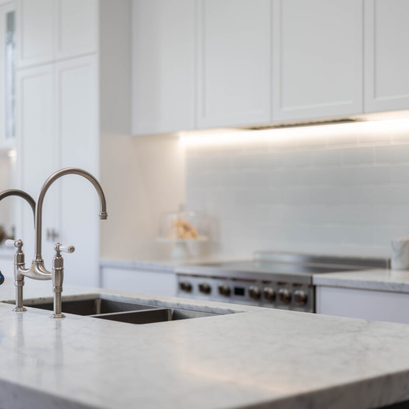 The kitchen style is classic Hamptons - shaker profile cabinetry, Carrara marble benchtops, with a soft blue-grey vein against a white base, nickel hardware, and a freestanding cooker. White subway tiles keep the back run of the space light and uniform. The Island The mid tone neutral grey of the island provides a depth of colour between the light and bright white cabinetry, and traditional warm toned parquetry. Entertainer's Delight The core kitchen space is balanced and mirror image. Extending down towards the windows, the kitchen transforms into an entertaining space; with a glass door cabinet, wine fridge, and space for four stools, mimicking a traditional table setting, with two either side.