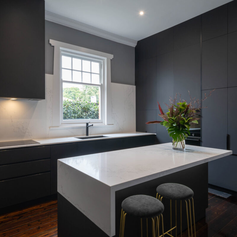 Dark and dramatic Kitchen. Black touch cabinetry with a white marble style benchtop on the island and the back run of cooking space. Large window letting in light and space. Sleek and modern.