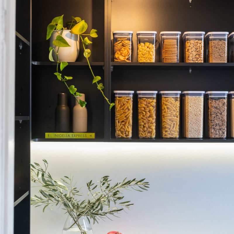 Touch Cabinetry. Black walk in pantry with white stone benchtop. Open shelving with containers and plants.