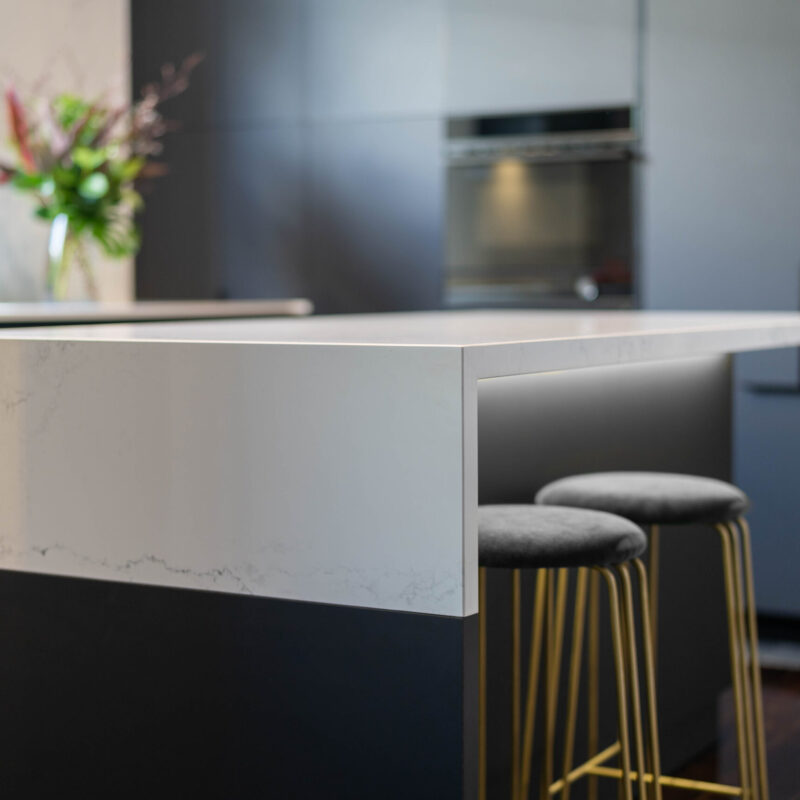 Waterfall end grey and white tones stone benchtop with under led lighting, black touch cabinetry and gold stools.