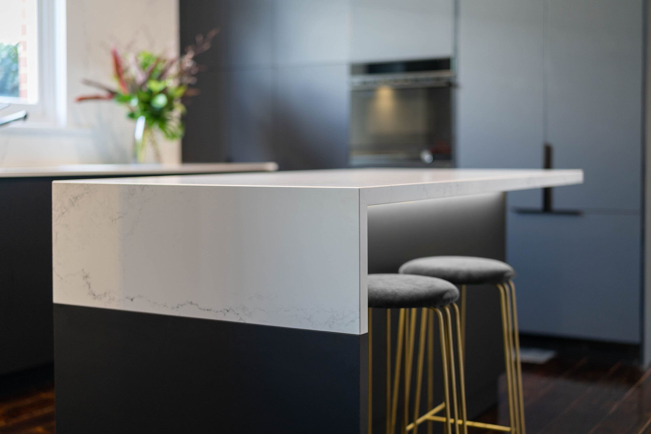 Waterfall end grey and white tones stone benchtop with under led lighting, black touch cabinetry and gold stools.