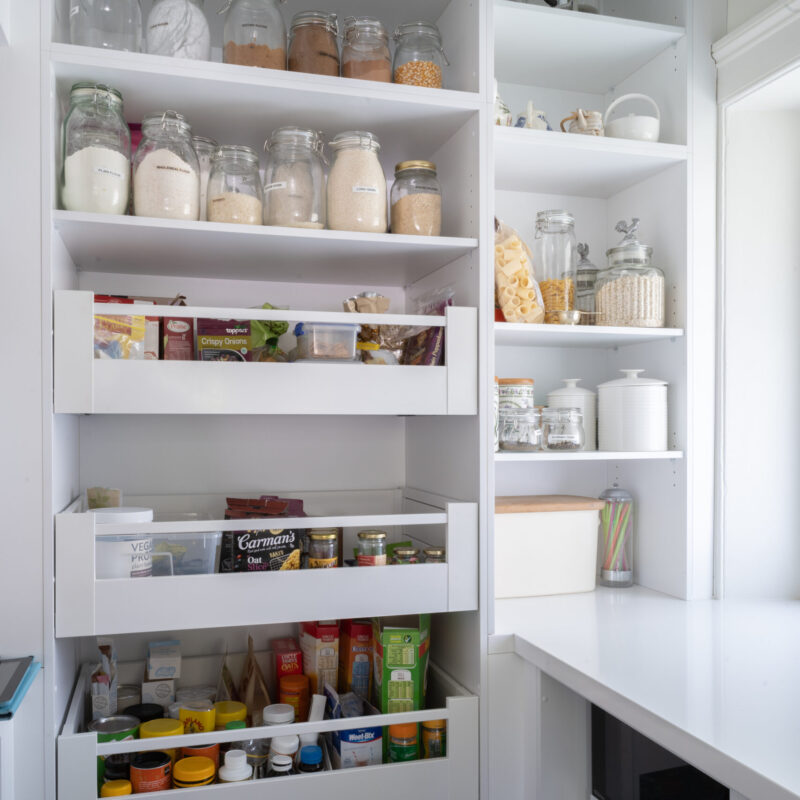 Walk in pantry, white and light full of pull out see through draws and cabinet space storage. White stone benchtop housing a microwave hidden in the base cabinets.