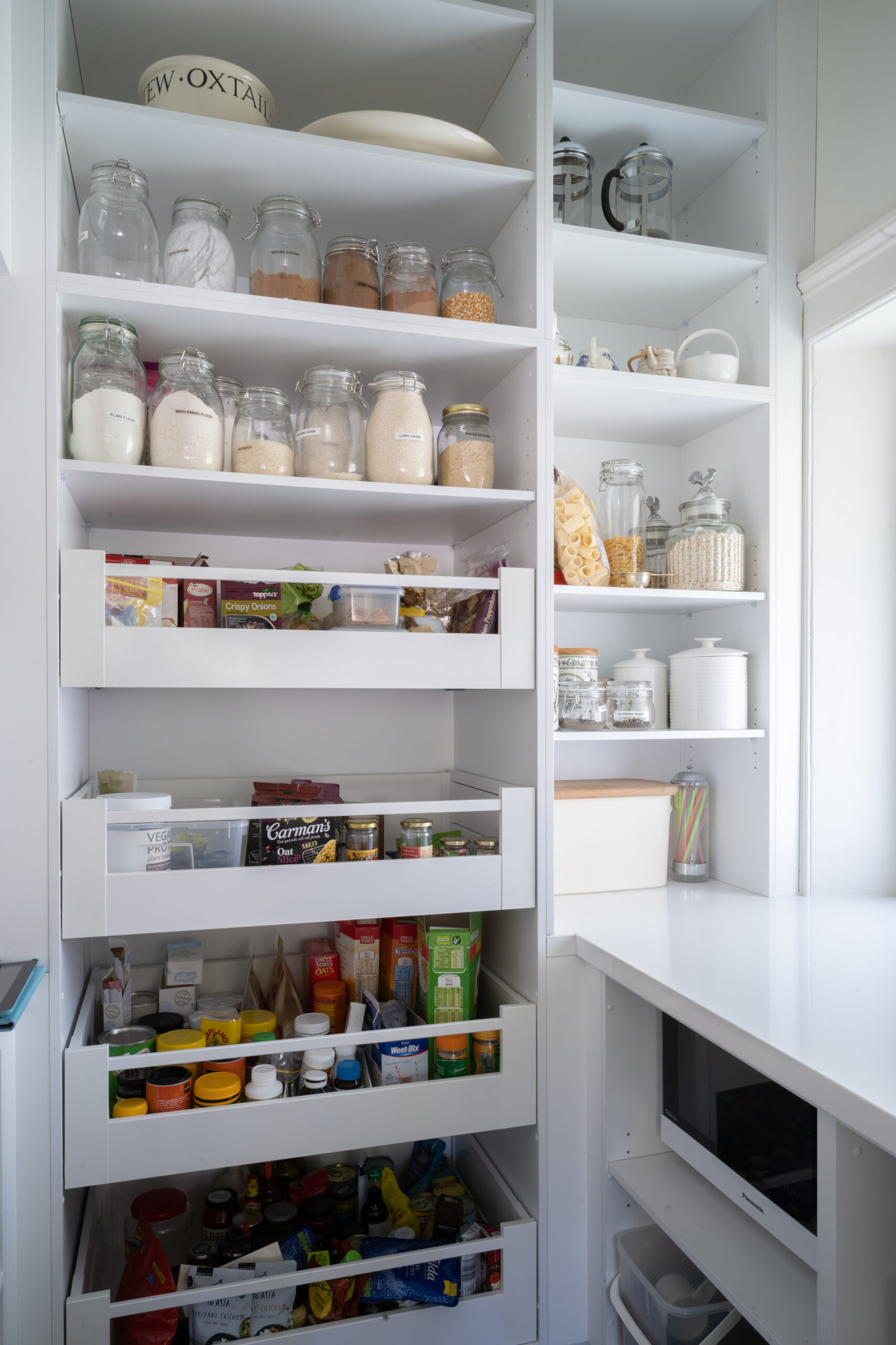 Walk in pantry, white and light full of pull out see through draws and cabinet space storage. White stone benchtop housing a microwave hidden in the base cabinets.