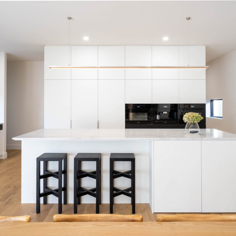 Stylish and modern Kitchen. Open plan light and white. Premium white TOUCH™ super matte cabinetry, anchored by a natural palette of blonde timber flooring, concrete paneling and marble benchtops, together juxtaposed by striking black highlights.