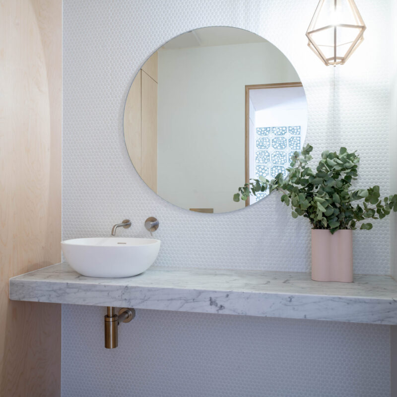 Beautiful powder room space. Vanity unit with a thick benchtop and white basin with small tiled splashback and silver, blond timber and gold highlights.