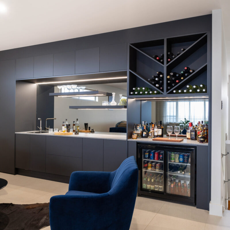 A modern home bar with black touch seamless cabinetry housing storage, shelving, fridges and wine racks with mirror and strip lighting.