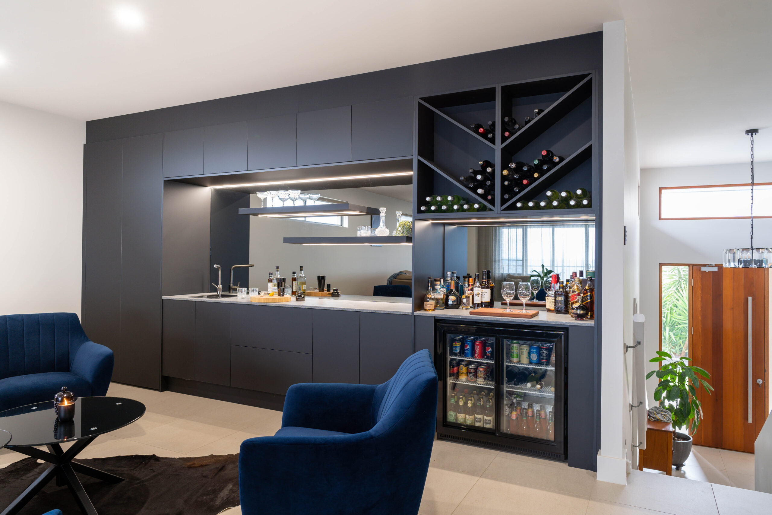 A modern home bar with black touch seamless cabinetry housing storage, shelving, fridges and wine racks with mirror and strip lighting.