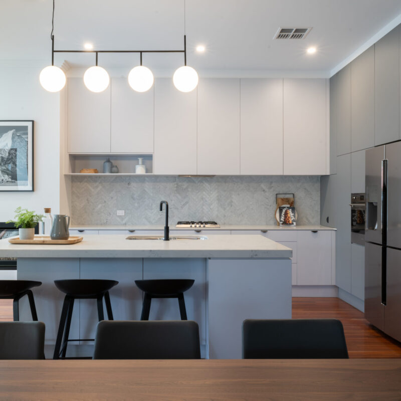 Open plan living. Modern Art deco Kitchen. Grey tones with white grey cabinetry, black cup handles and black highlights. Herringbone grey marble splashback and timber floors are added with the darker grey cabinetry on the side to house the appliances.