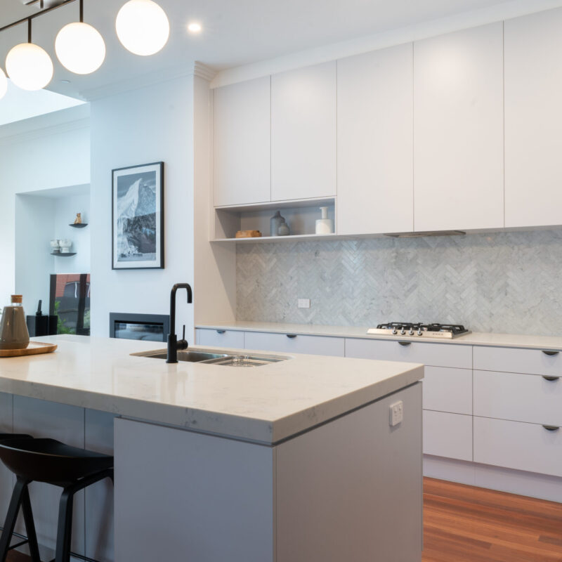 Modern Art deco Kitchen. Grey tones with white grey cabinetry, black cup handles and black highlights. Herringbone grey marble splashback and timber floors are added.