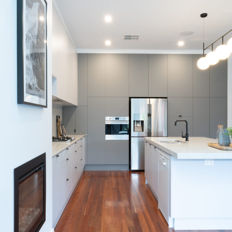 Modern Art deco Kitchen. Grey tones with white grey cabinetry, black cup handles and black highlights. Herringbone grey marble splashback and timber floors are added with the darker grey cabinetry on the side to house the appliances.