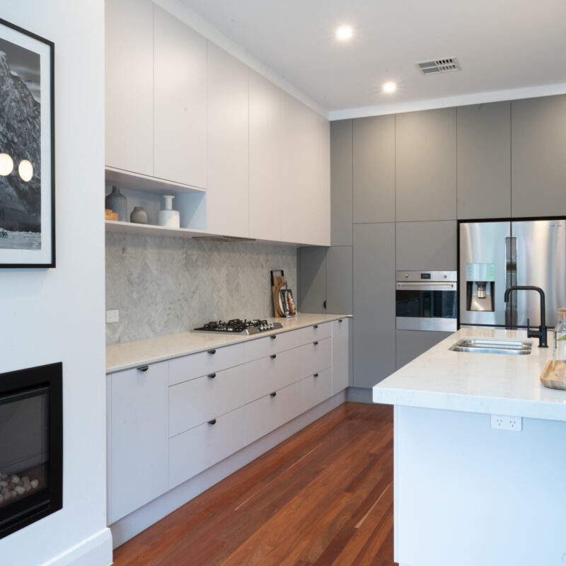 Modern Art deco Kitchen. Grey tones with white grey cabinetry, black cup handles and black highlights. Herringbone grey marble splashback and timber floors are added with the darker grey cabinetry on the side to house the appliances.