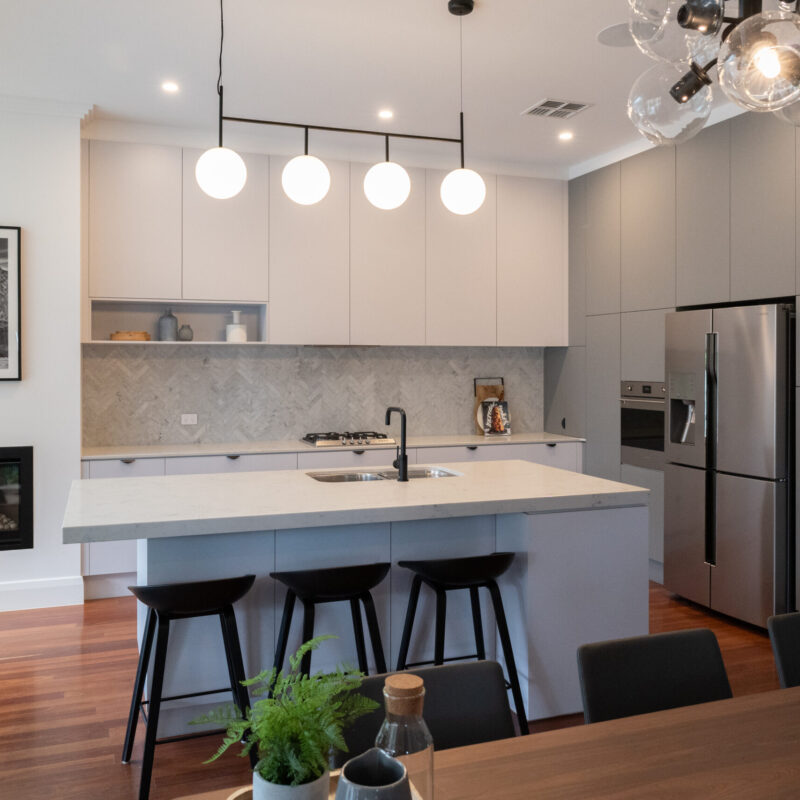 Modern Art deco Kitchen. Grey tones with white grey cabinetry, black cup handles and black highlights. Herringbone grey marble splashback and timber floors are added with the darker grey cabinetry on the side to house the appliances. Large open space.