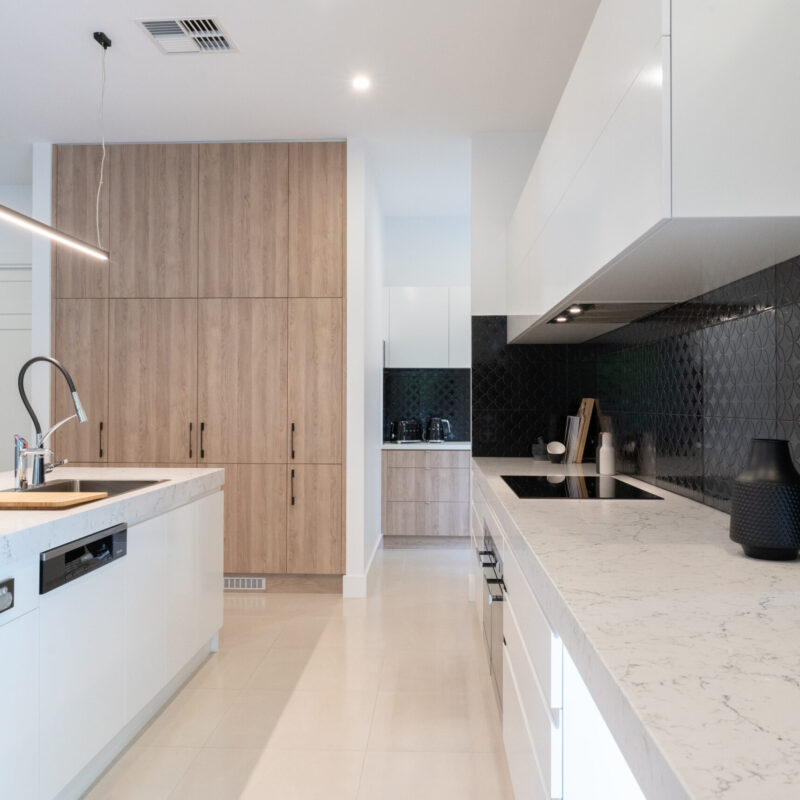 A modern masterpiece kitchen. Thick stone benchtops in grey tones with black, white and timber features. Large island with integrated dishwasher and large sink and tap.