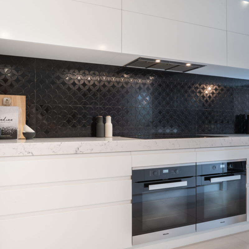 A modern kitchen cooking space. white cabinetry, black patterned gloss splashback, white draw banks, overheads and Miele ovens and undermount rangehood.