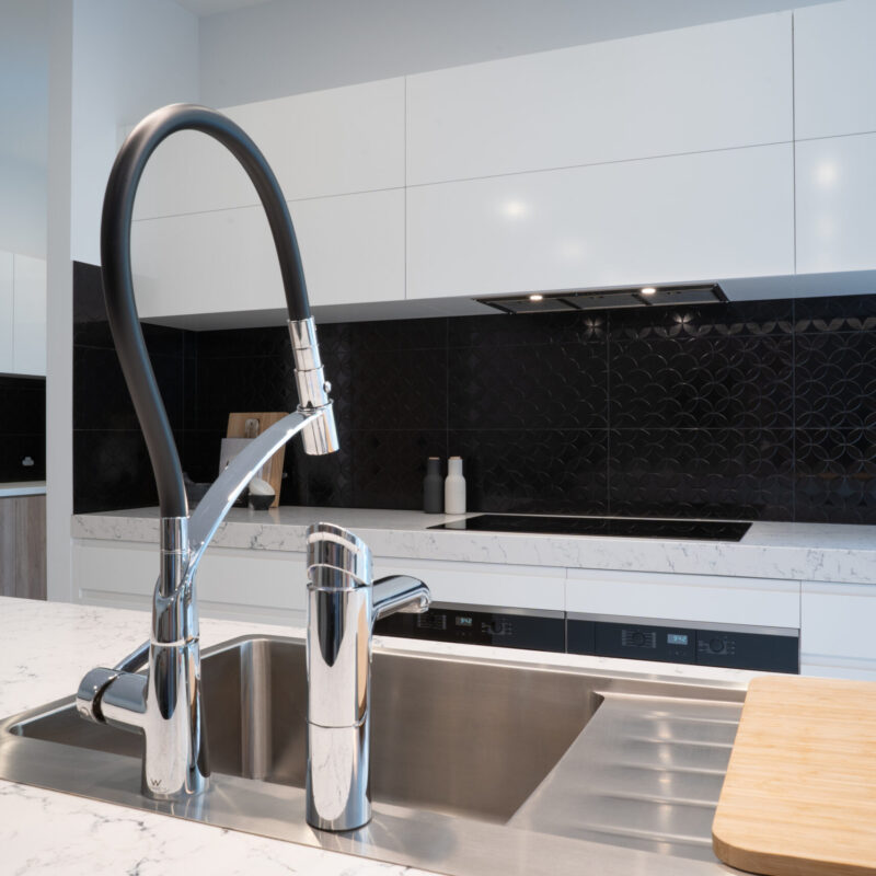 A modern kitchen cooking space. white cabinetry, black patterned gloss splashback, white draw banks with Oliveri stainless steel sink and taps.