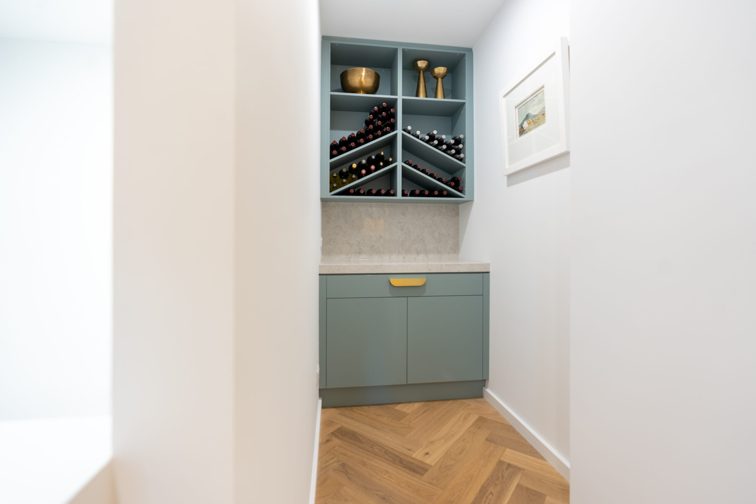 Small butlers in Fjord. Stone benchtop and matching stone splashback. Brushed gold handles and an open shelf wine rack overhead cabinet.