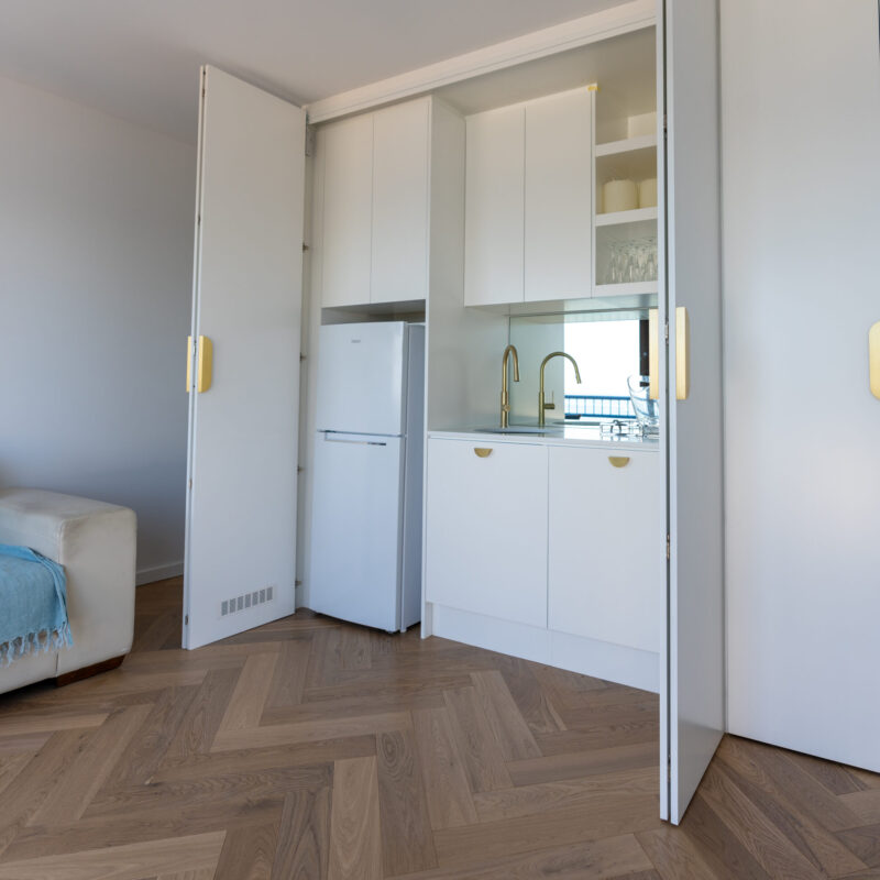 Modern kitchenette hidden behind white full height bi-fold doors. White kitchens cabinets with a white stone benchtop and mirror splashback. Brushed gold tapware and handles.
