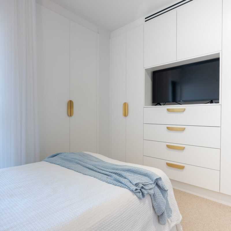 Bedroom Wardrobe. White floor to ceiling cabinets. An open cabinet in the middle to house a television with drawers below and overhead cabinets above. Brushed gold handles.