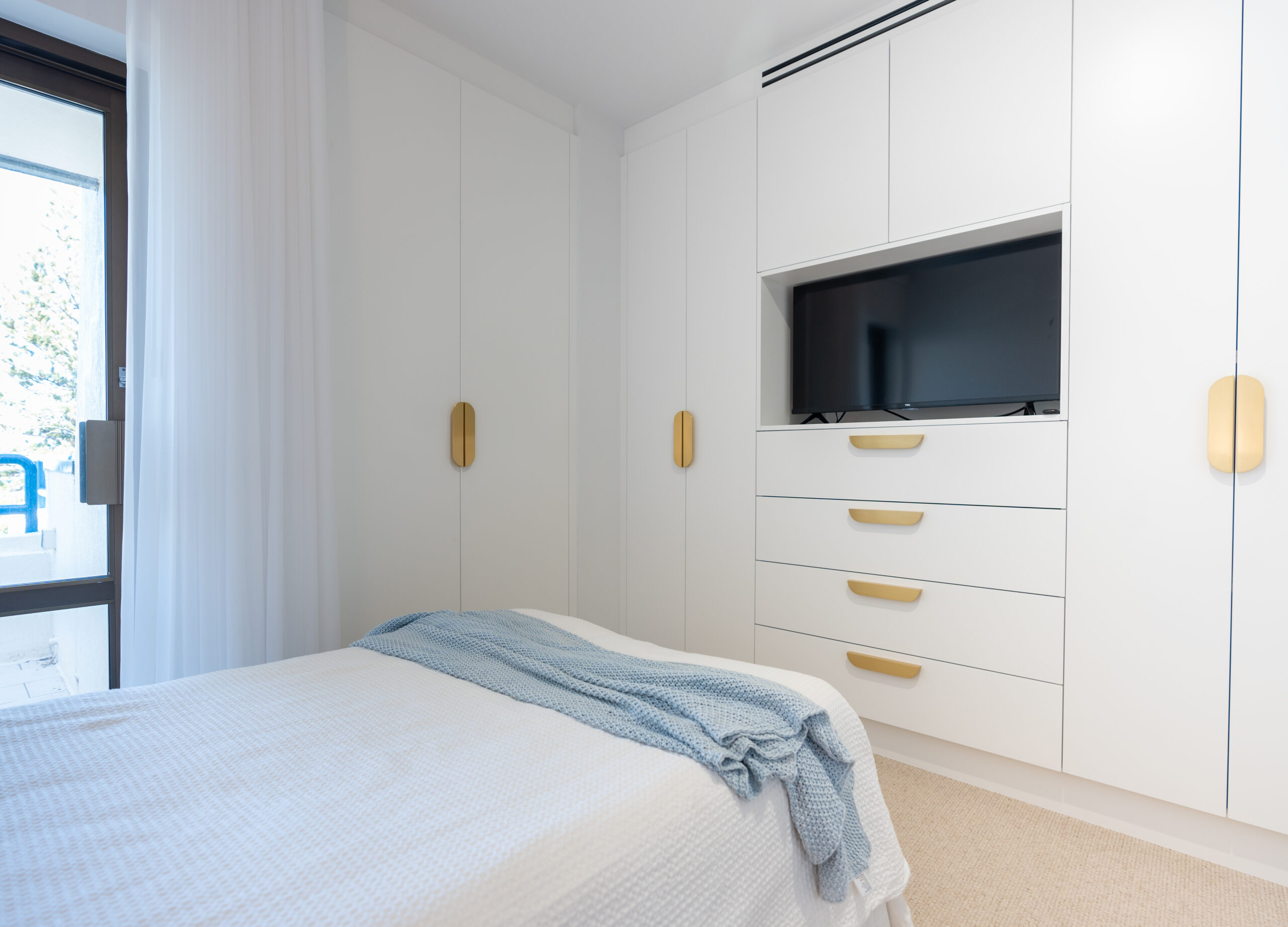 Bedroom Wardrobe. White floor to ceiling cabinets. An open cabinet in the middle to house a television with drawers below and overhead cabinets above. Brushed gold handles.