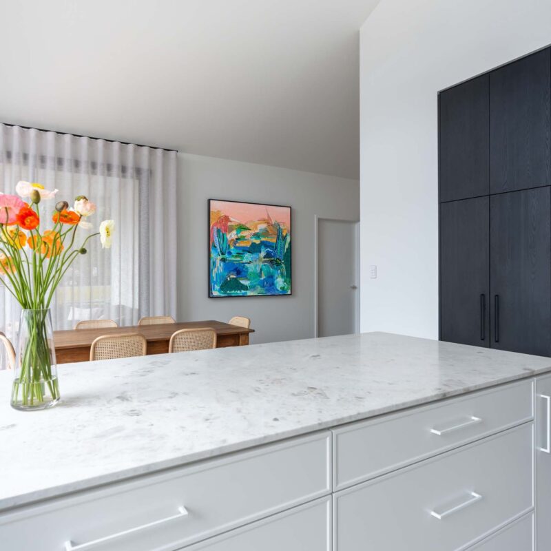 Island bench featuring slim shaker style profiled white cabinets and a light marble island benchtop. To the right are floor to ceiling cabinets in a black wood grain, including built in appliances.