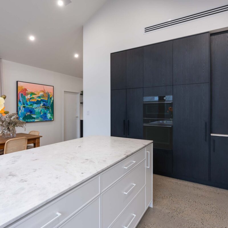 Island bench featuring slim shaker style profiled white cabinets and a light marble island benchtop. To the right are floor to ceiling cabinets in a black wood grain, including built in appliances.