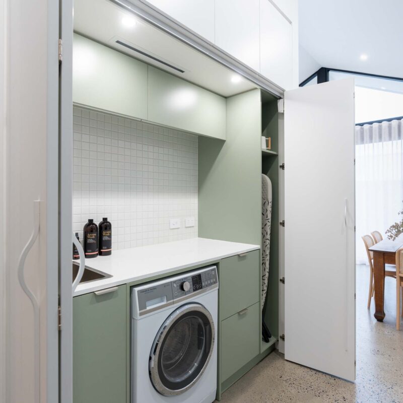 Matte green concealed laundry with a light stone benchtop and small square tile splashback. Includes an under-bench washing machine and broom cabinet, all hidden behind matte white sliding doors.