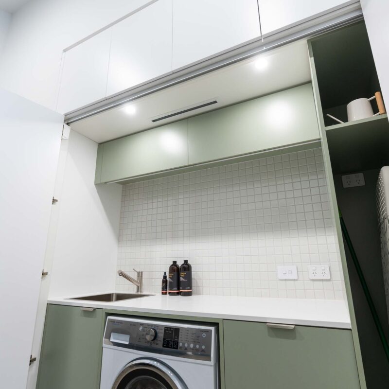 Matte green concealed laundry with a light stone benchtop and small square tile splashback. Includes an under-bench washing machine and broom cabinet, all hidden behind matte white sliding doors.