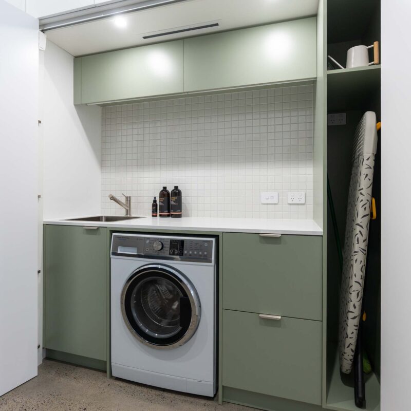 Matte green concealed laundry with a light stone benchtop and small square tile splashback. Includes an under-bench washing machine and broom cabinet, all hidden behind matte white sliding doors.