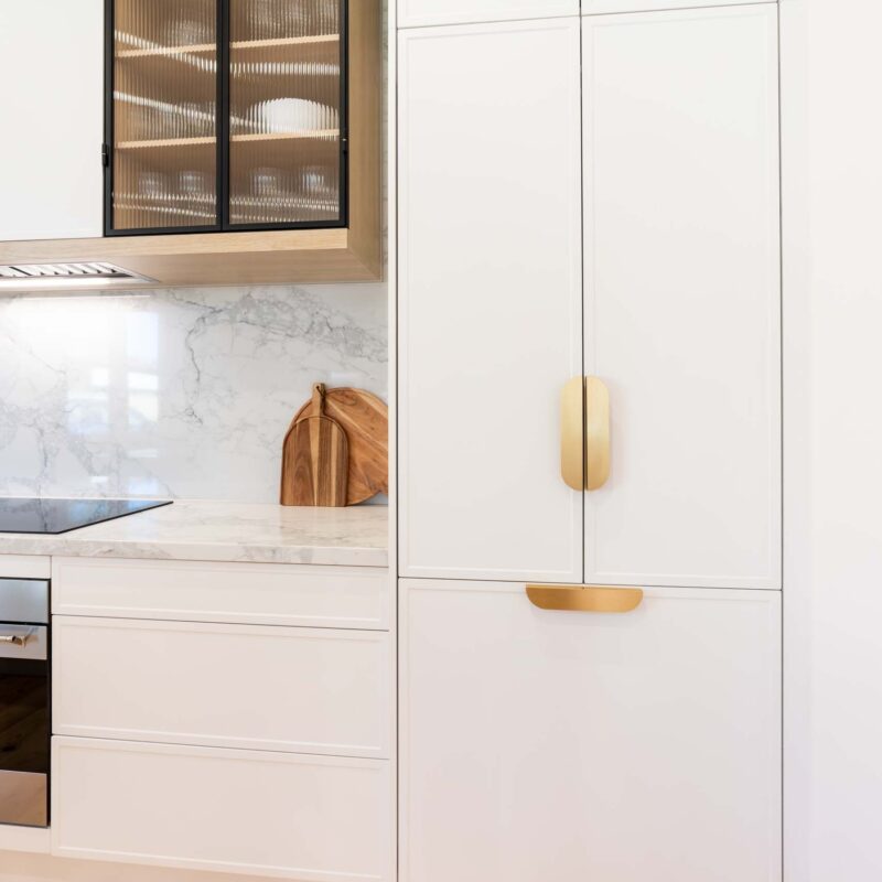 White slim shaker-profile doors conceal a fridge-freezer, finished with matte gold handles. A light marble benchtop sits alongside white slim shaker cabinetry on the left.