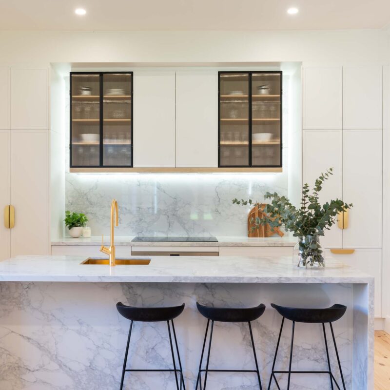 White kitchen and island bench featuring white slim shaker profile doors and drawers and light marble benchtops. Glass fluted overhead cabinets. Brushed gold handles.