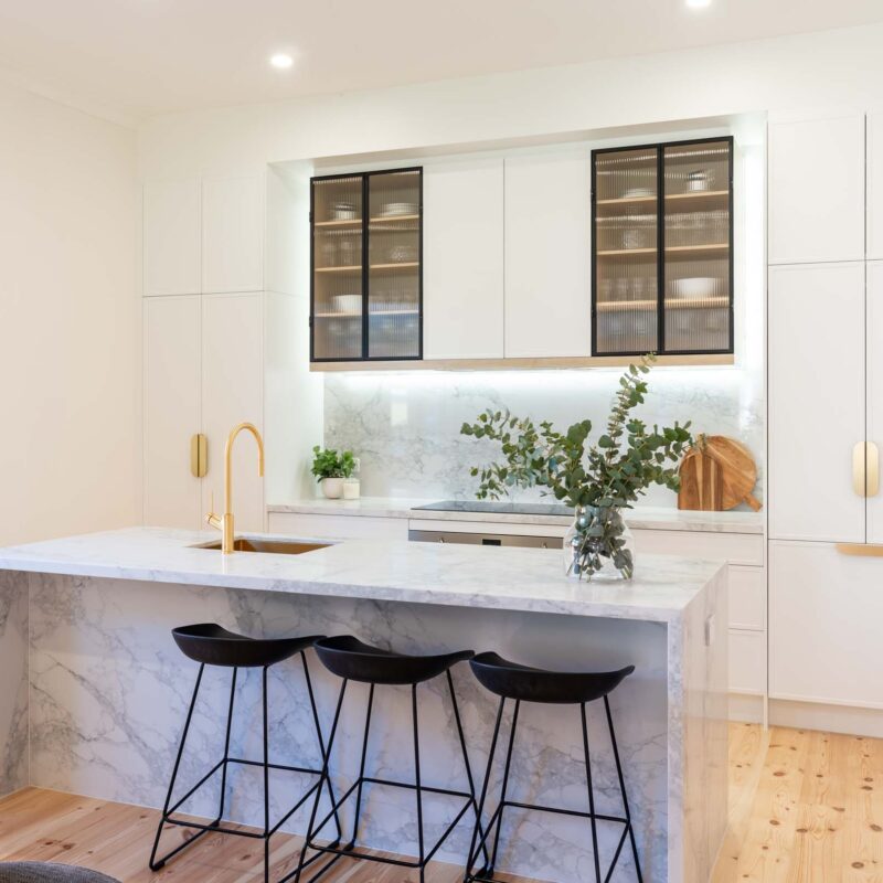 White kitchen and island bench featuring white slim shaker profile doors and drawers and light marble benchtops. Glass fluted overhead cabinets. Brushed gold handles.