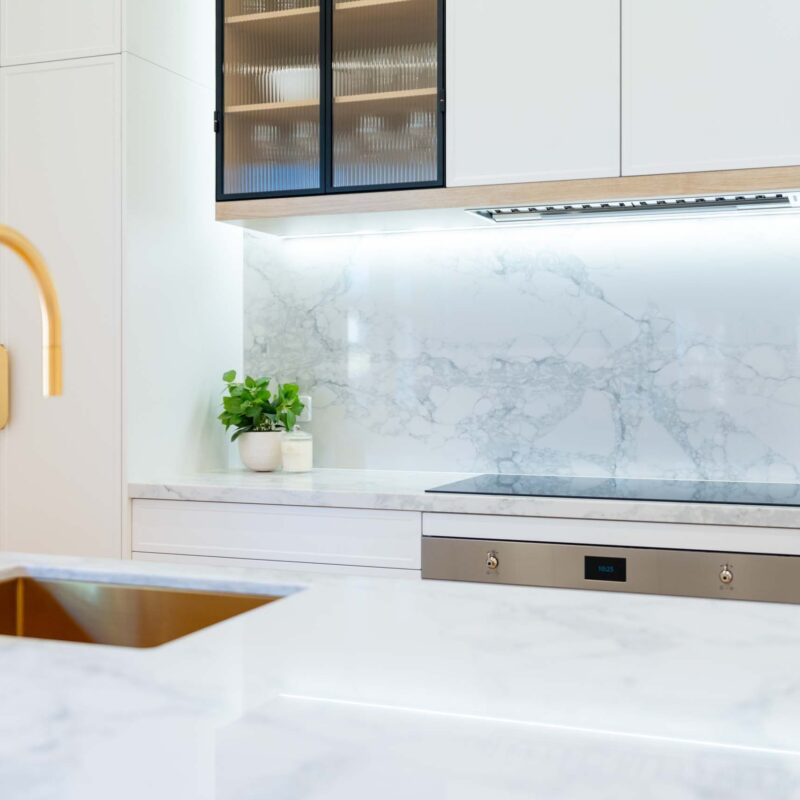 White galley style kitchen featuring white slim shaker profile doors and drawers and light marble benchtops. Glass fluted overhead cabinets. Brushed gold handles.