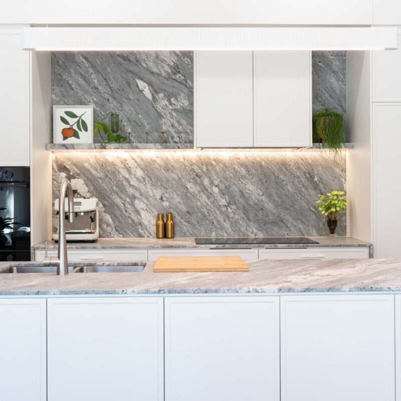 Modern kitchen with large island benchtop. White thin shaker profiled cabinets with a bold grey marble benchtop and splashback. Large concealed double door fridge with matt white handles.